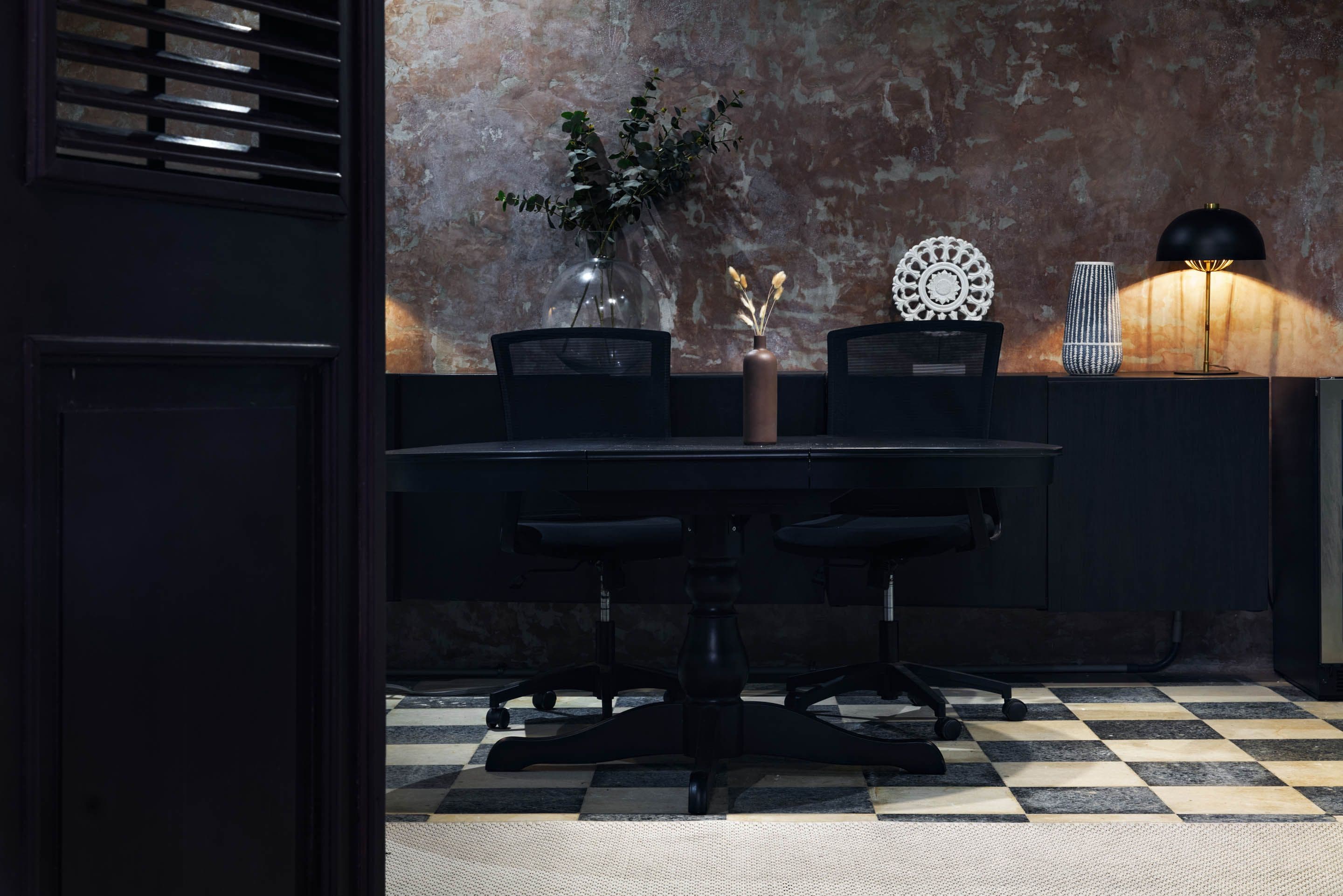 Un área de comedor con piso a cuadros, una mesa con sillas, un jarrón con plantas secas, un plato decorativo y una lámpara.