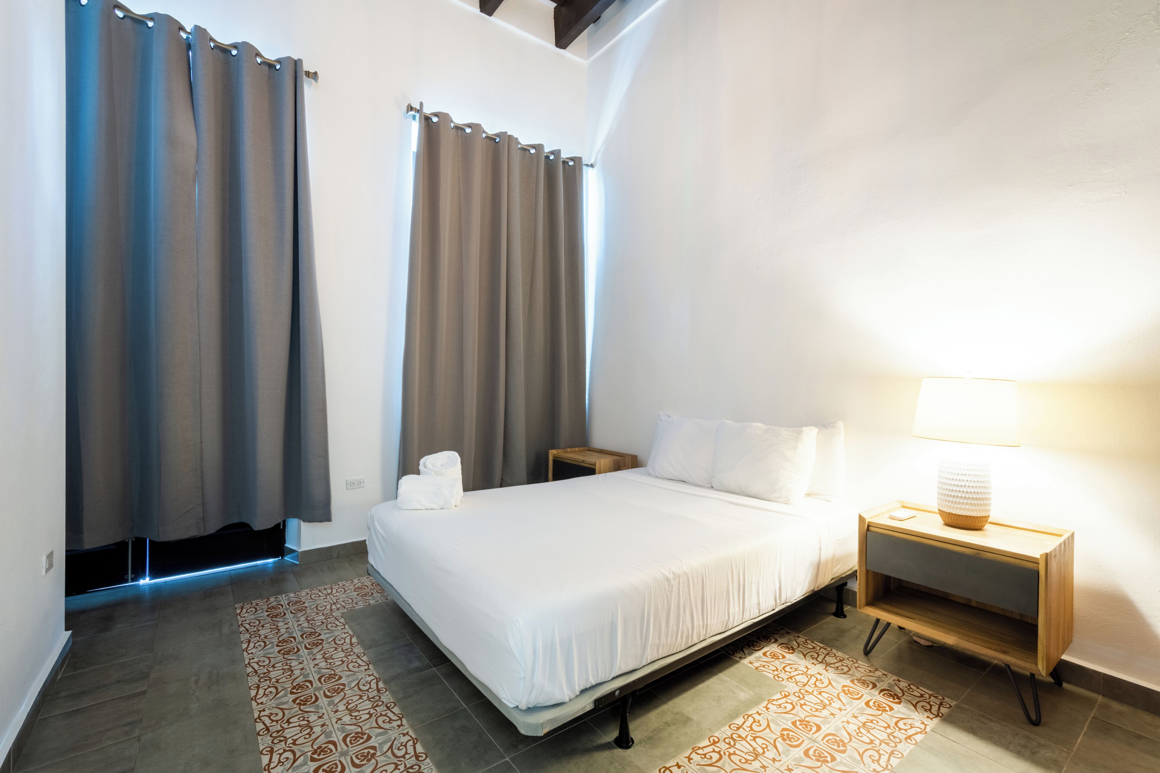 A hotel room with a bed, a nightstand, a lamp, and a view of the outside.