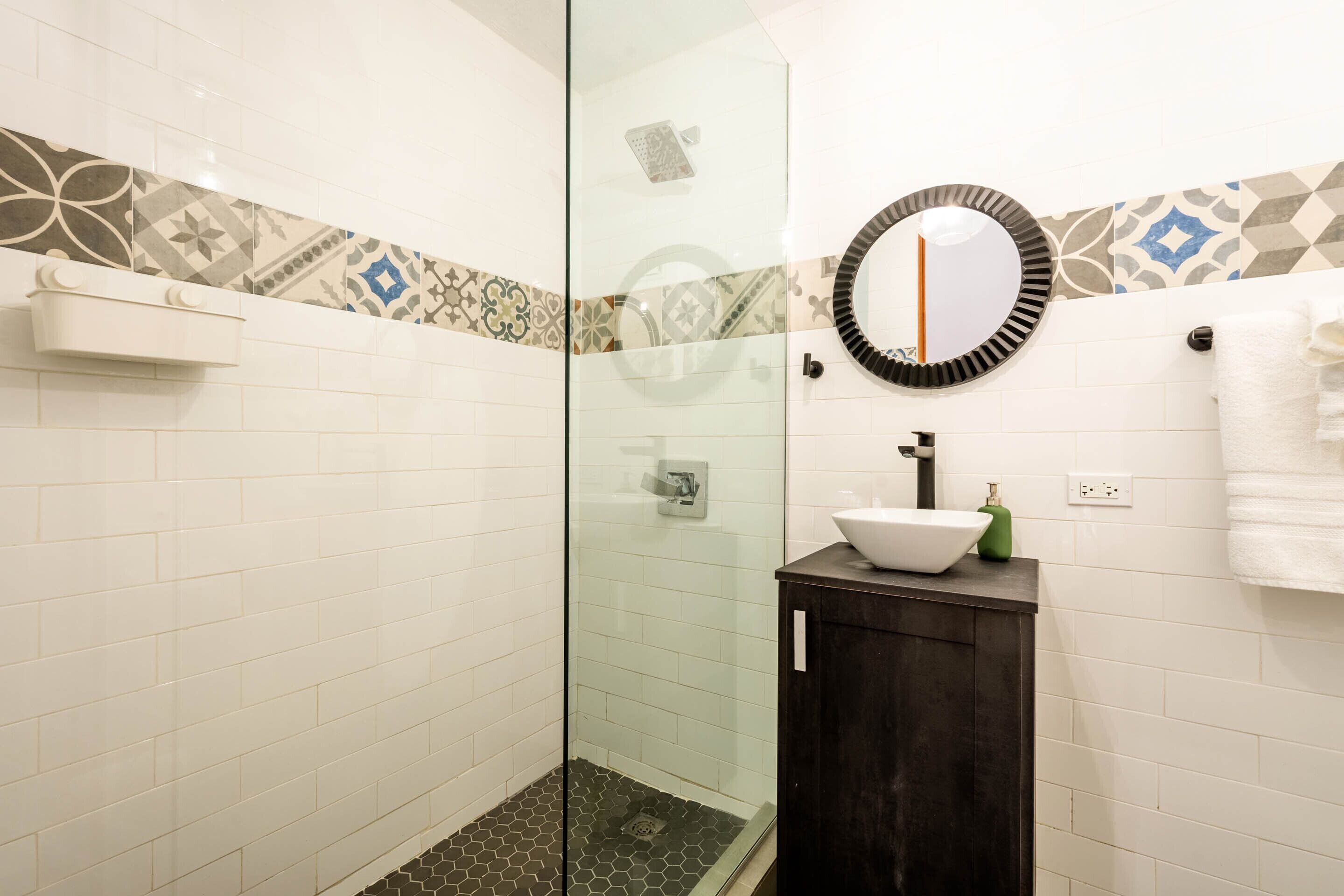 A bathroom with a glass shower enclosure, a white sink with a dark cabinet, and patterned tiles on the walls.