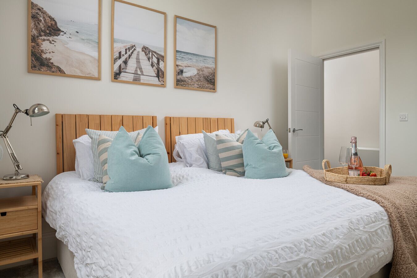 A neatly arranged bedroom with a wooden headboard, a bed with white bedding and light blue pillows, a bedside table with a lamp, and a nightstand. There are two framed pictures on the wall above the bed.