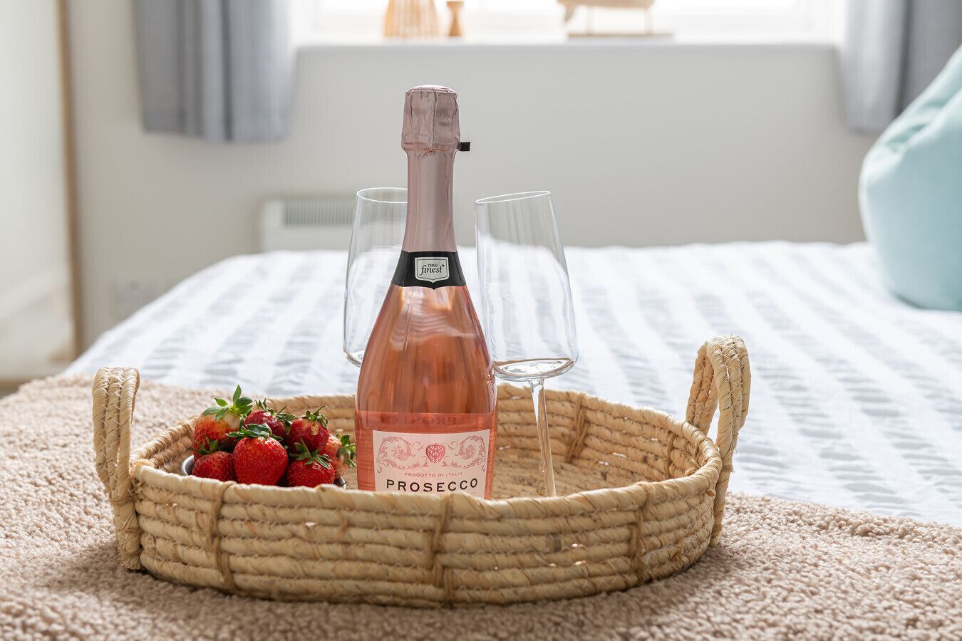 A wicker basket containing a bottle of prosecco, two glasses, and a bowl of strawberries on a bed with a striped blanket.