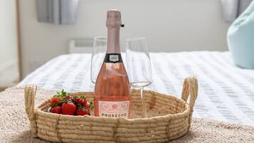 A wicker basket containing a bottle of prosecco, two glasses, and a bowl of strawberries on a bed with a striped blanket.
