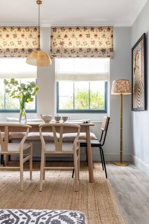 Un coin repas comprenant une table, des chaises, un vase avec des fleurs, une lampe et une fenêtre agrémentée de rideaux à motifs floraux.