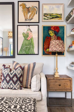 Una sala de estar con un sofá, almohadas, una mesita de noche de madera con una lámpara y obras de arte enmarcadas en la pared.