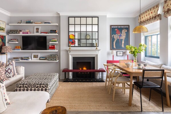 A living room with a fireplace, a television, a dining area with chairs, and a bookshelf with books. - William Mews by onefinestay (London)