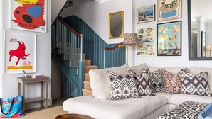 A living room with a staircase, a sofa with patterned cushions, and various framed artworks on the walls.