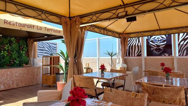 An outdoor restaurant with tables and chairs, a sign reading "Restaurant La Terrasse", and a view of the sky through large windows. - ZEBRA RESIDENCE (Bambylor)