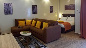 A modern living room with a brown sofa, a round coffee table, and a bed with an orange blanket. There are ceiling fans, wall art, and a patterned rug. - ZEBRA RESIDENCE (Bambylor)