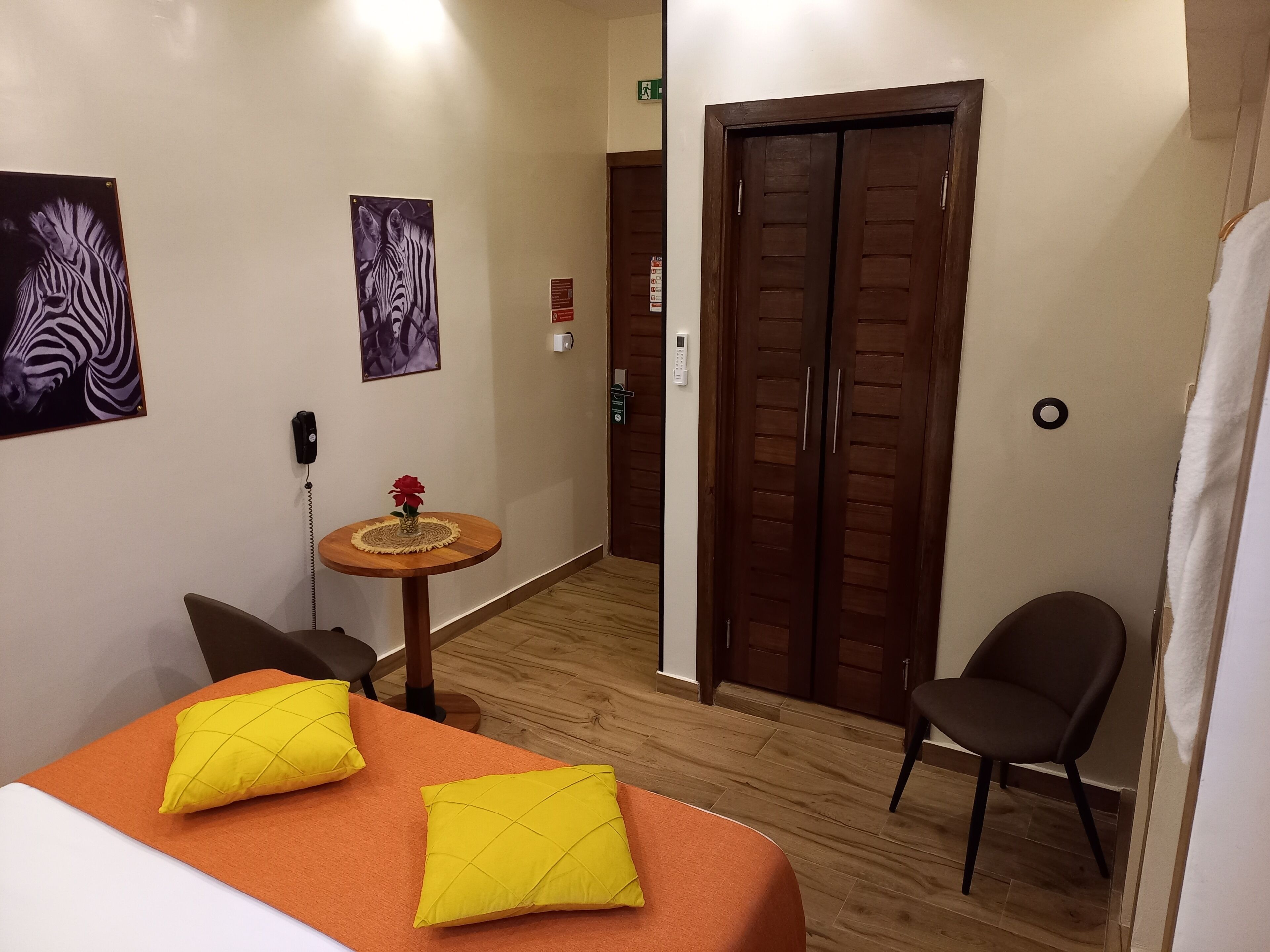 Een hotelkamer met een bed, twee gele kussens, een klein tafeltje met een bloemenvaas, een stoel en een houten vloer.