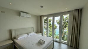 A bedroom with a bed, a nightstand, an air conditioning unit, and a view of the ocean through large sliding doors.