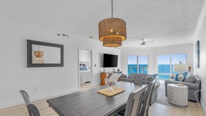 A spacious living room with a dark wooden dining table, grey sofas, and a large window offering a view of the ocean. - NEW 15th Floor Gulf View - Balcony - Pool - Hot Tub (Pensacola Beach)
