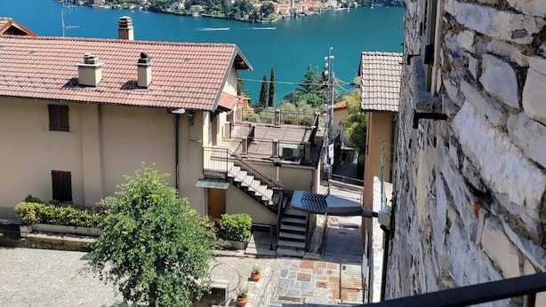 A view of a lake with a mountain in the background, a stone building with a balcony, and a small courtyard with steps leading to the building.
