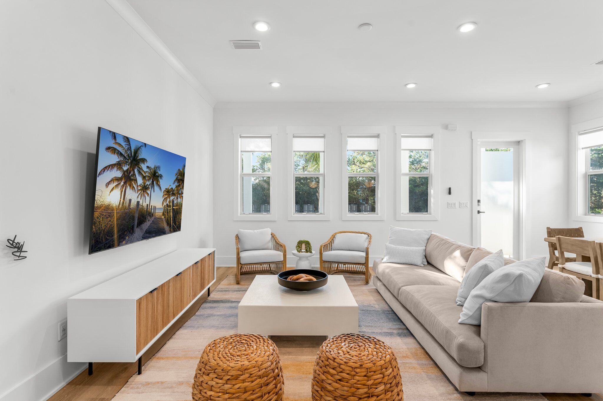 A modern living room with a white color scheme, a large flat-screen TV mounted on the wall, a beige sofa, two wicker ottomans, and a white coffee table with a bowl of fruit.