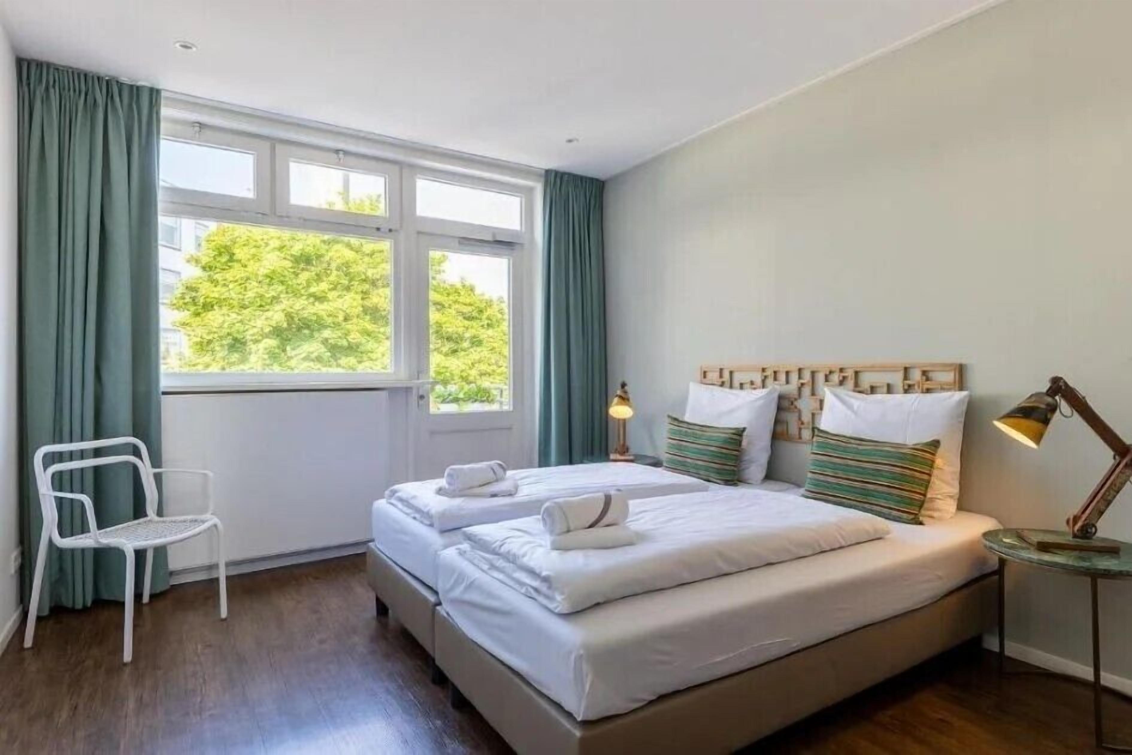 A hotel room with two beds, a chair, a lamp, and a view of greenery outside.