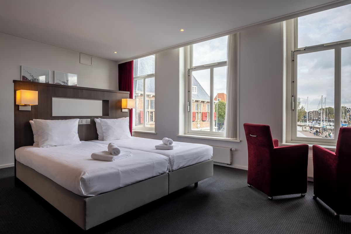 Habitación de hotel moderna con una cama grande, dos mesitas de noche, un escritorio y una silla. Amplias ventanas ofrecen vistas a la ciudad.