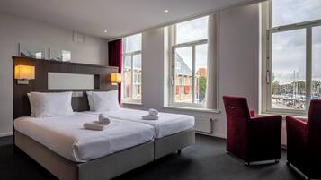 Quarto de hotel moderno com uma cama grande, duas mesas de cabeceira, uma escrivaninha e uma cadeira. Janelas amplas oferecem vista para a cidade.