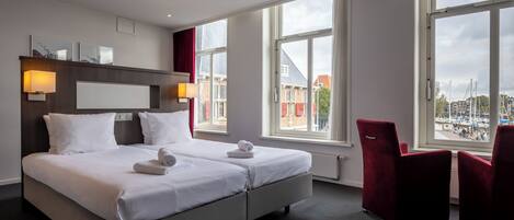 A modern hotel room with a large bed, two bedside tables, a desk, and a chair. There are large windows offering a view of the city.