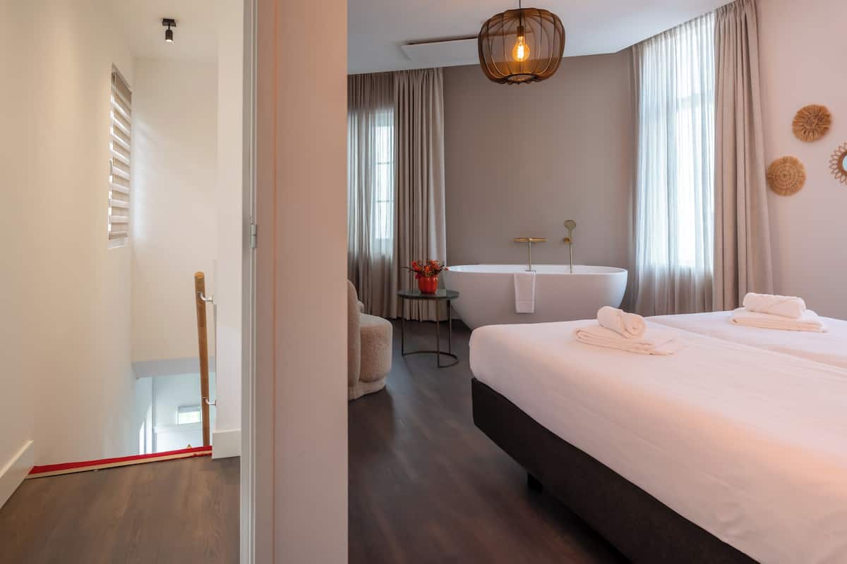 Habitación de hotel con una cama grande, un área de descanso con una silla y una mesita, una bañera y un candelabro en el techo.