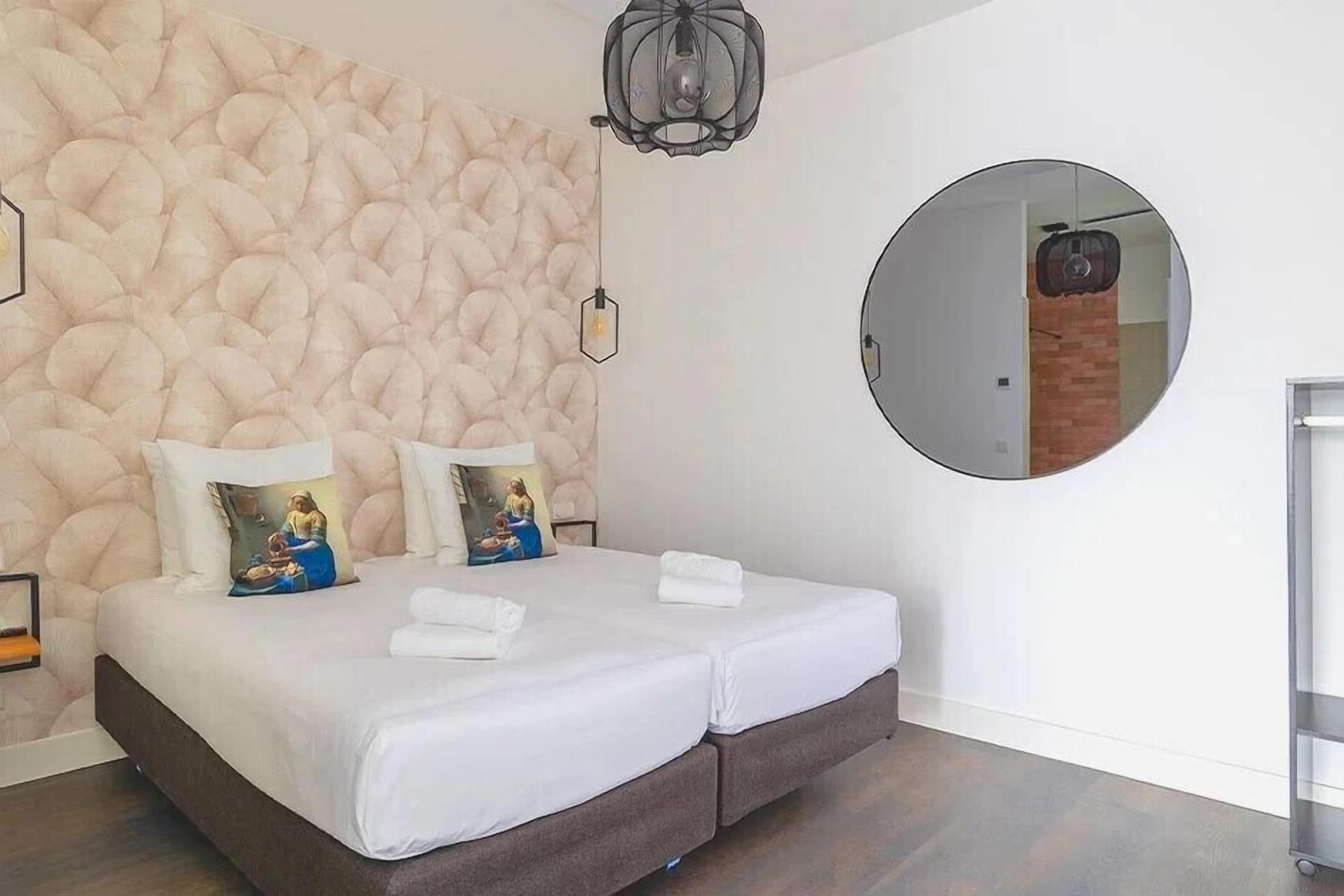 A double bed with white linens, a textured wall, a round mirror, and a hanging light fixture.