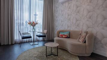 A modern living room with a beige sofa, two black chairs, a glass table, and a round coffee table. A decorative vase with flowers is on the table, and the room has a textured wall and a large window with sheer curtains.