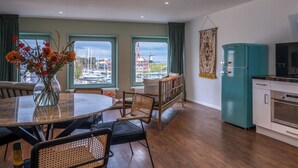 Une cuisine moderne avec un réfrigérateur de couleur turquoise, un coin repas comprenant une table et des chaises, et de grandes fenêtres offrant une vue sur un port de plaisance.