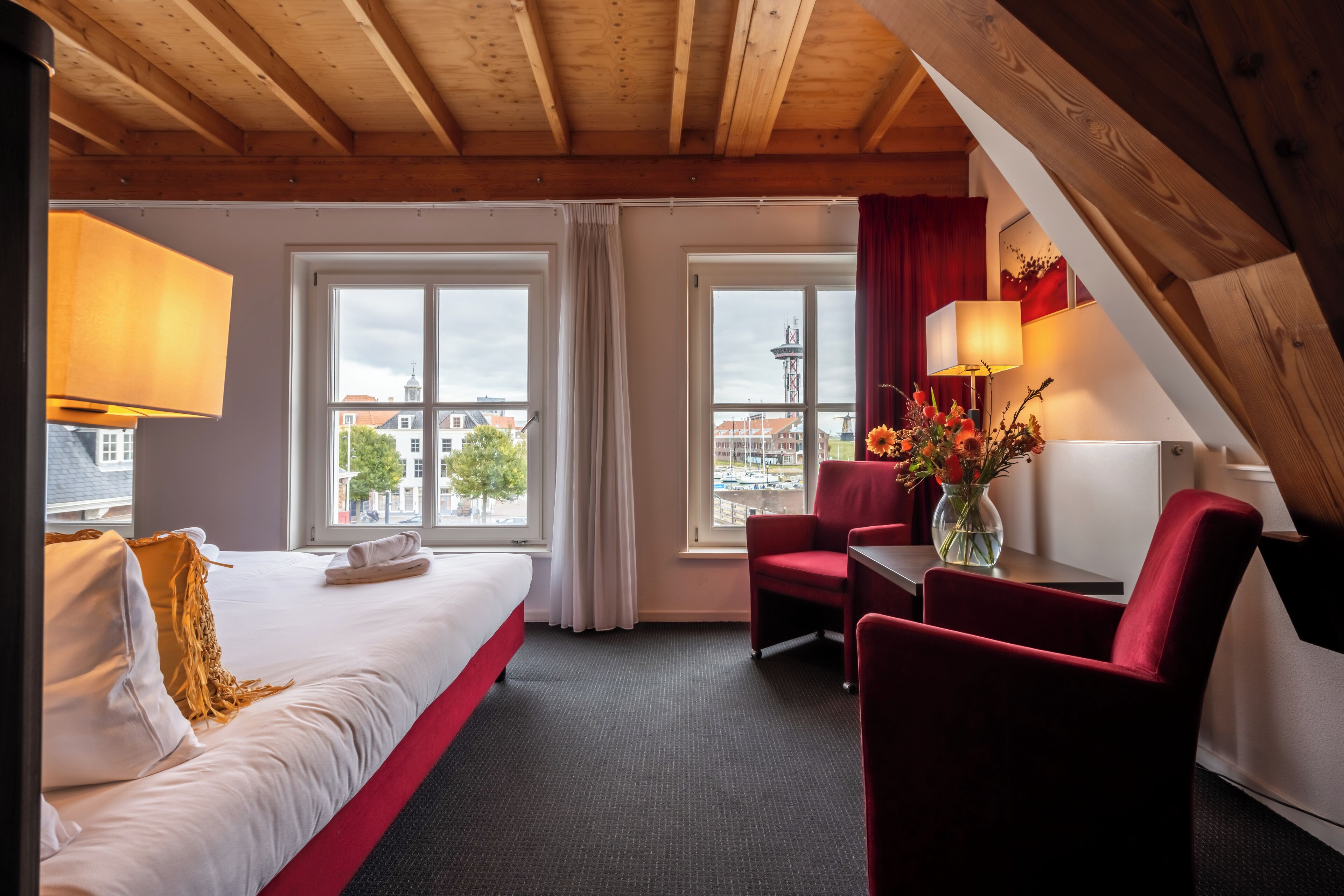 Een hotelkamer met een bed, twee ramen, een bureau met stoel en uitzicht op gebouwen.