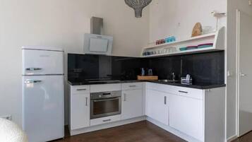 Cocina compacta con gabinetes blancos, horno integrado, microondas y refrigerador. Tiene una encimera negra y un lámpara colgante.