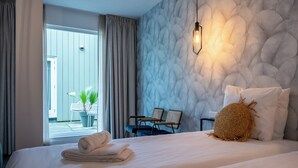 Une chambre d’hôtel avec un lit, deux chaises, une petite table, une plante en pot et une vue sur l’extérieur.