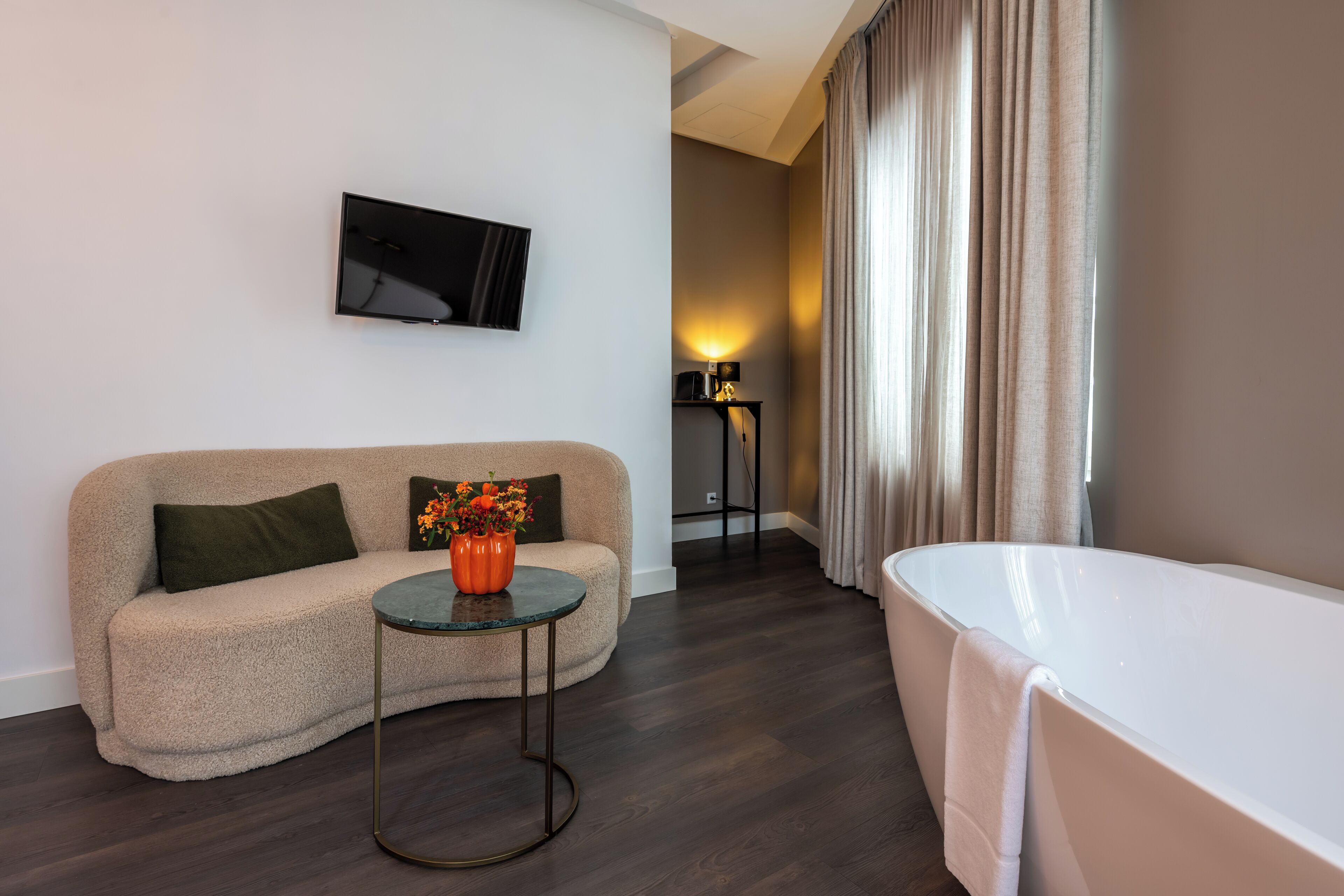 A modern hotel room with a beige sofa, a small round table with a vase of flowers, a flat-screen TV mounted on the wall, a standing lamp, and a white bathtub.