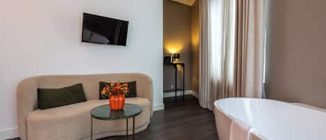 A modern hotel room with a beige sofa, a small round table with a vase of flowers, a flat-screen TV mounted on the wall, a standing lamp, and a white bathtub.