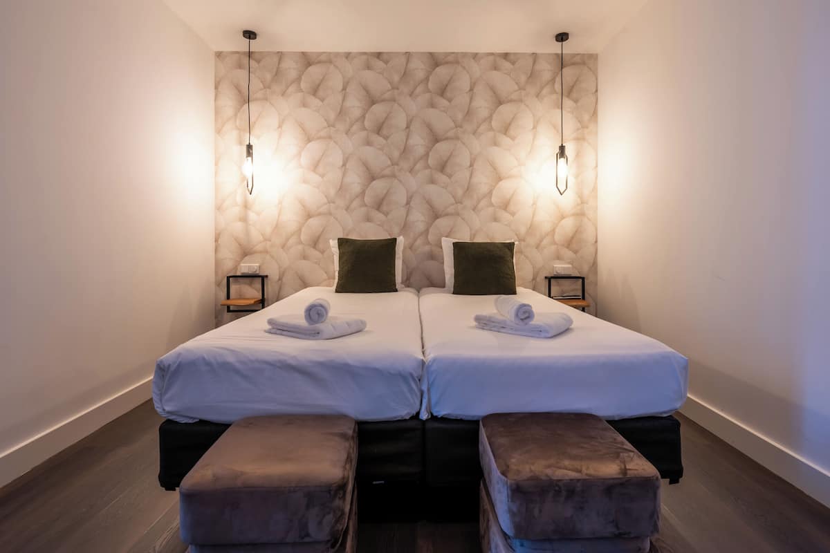 Habitación doble con dos camas, cada una con una almohada y una toalla, pared con diseño de piedra y dos mesitas de noche con lámparas.