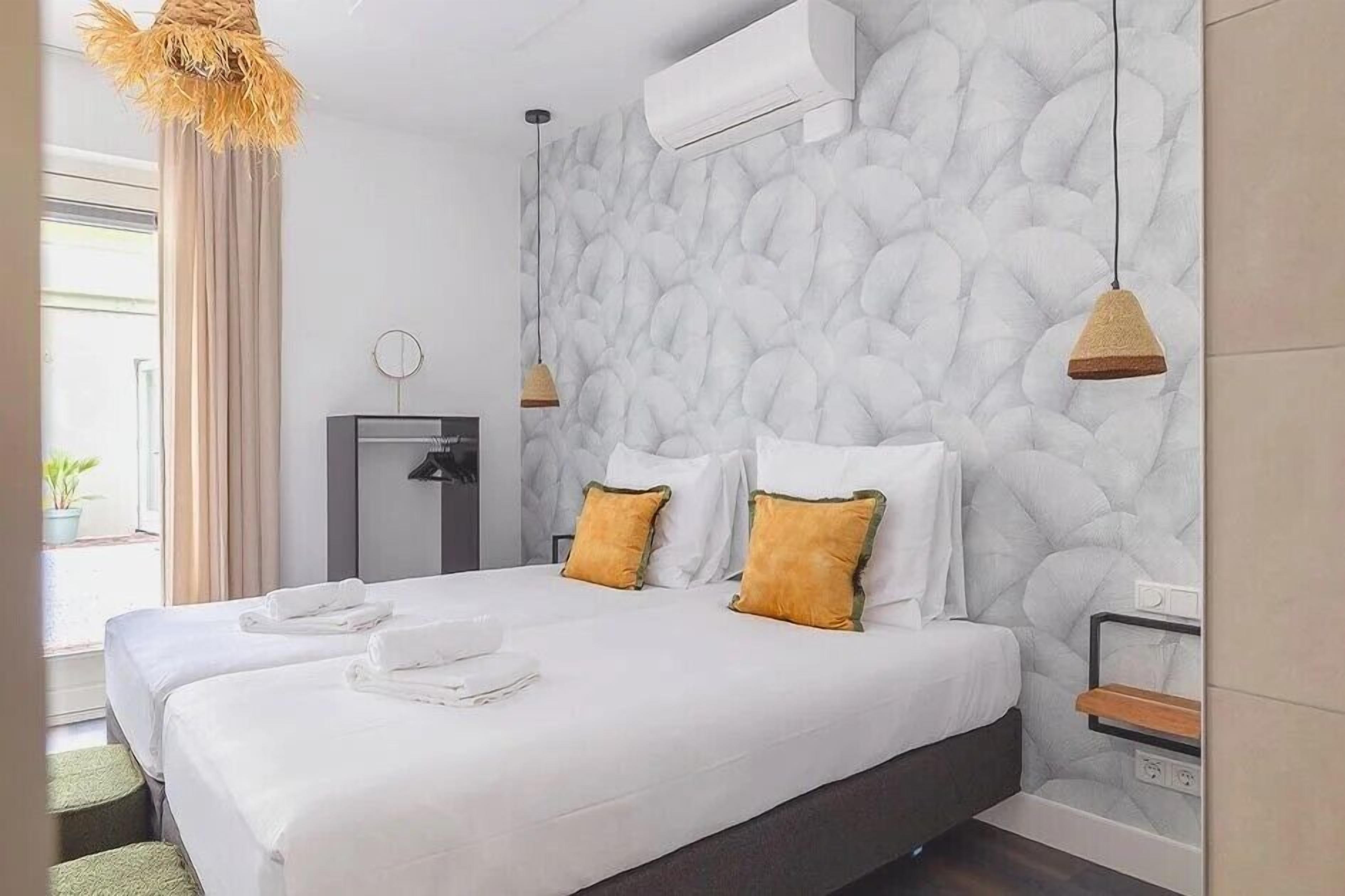 A hotel room with a large bed, two pillows, a bedside table, an air conditioning unit, and a window with curtains.