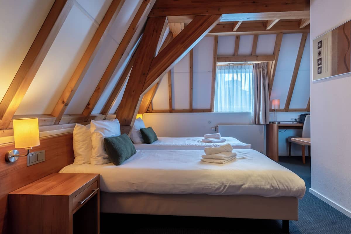 Una habitación con techo inclinado, vigas de madera, una cama con ropa blanca, una mesita de noche con lámpara y un escritorio con silla.