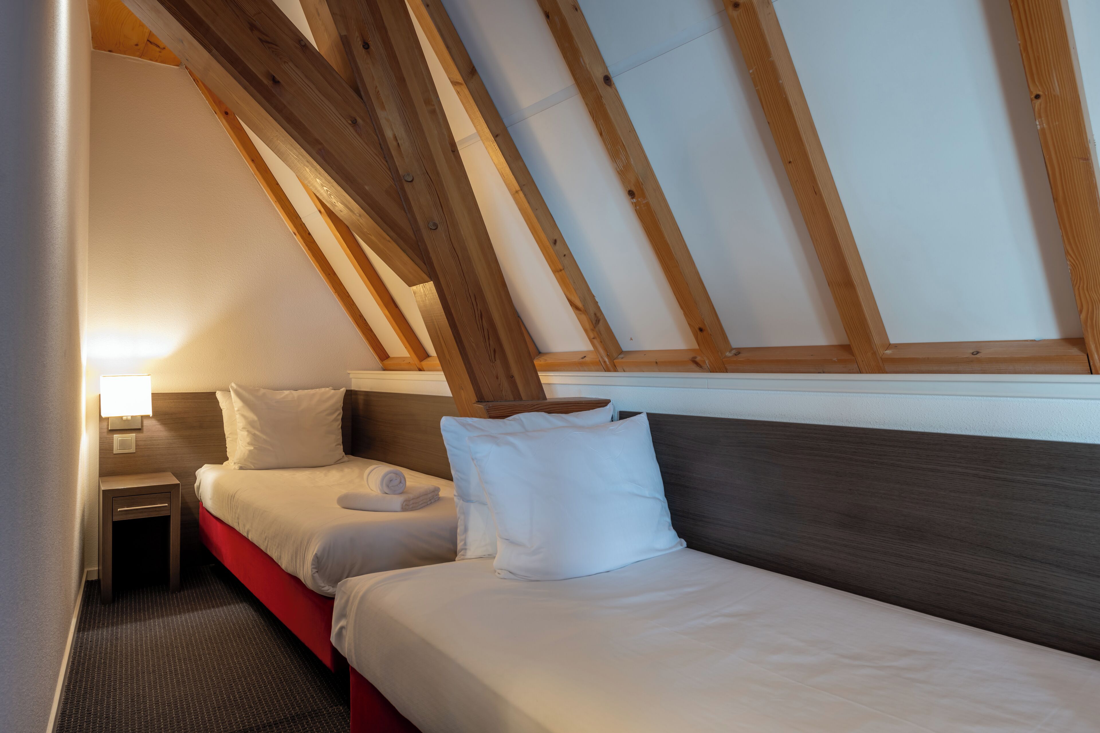 A hotel room with two beds, wooden beams, a nightstand, and a lamp.