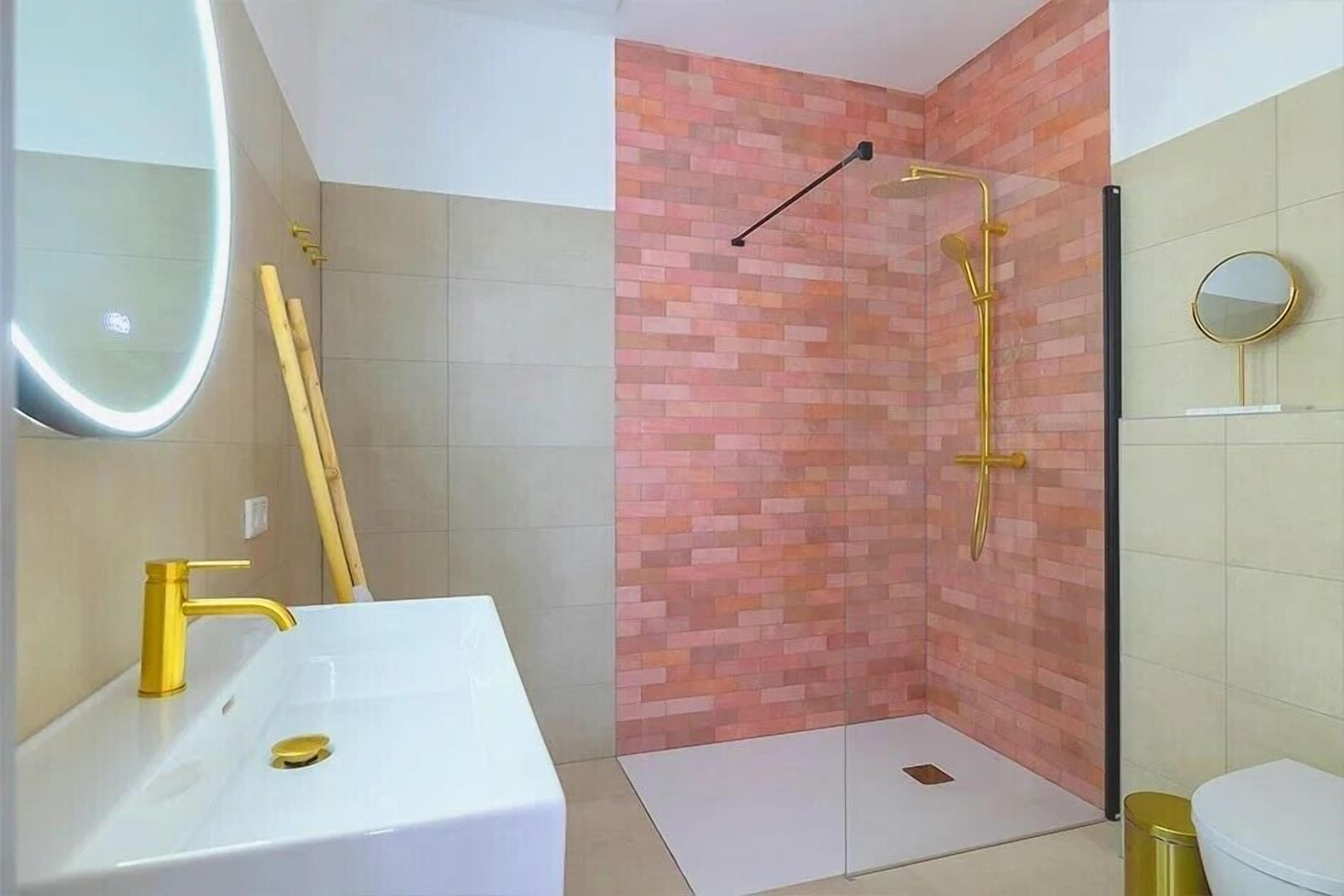 Une salle de bain avec un lavabo blanc, des robinets dorés, une douche avec un mur en briques et des toilettes.