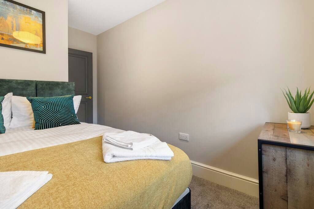 A neatly made bed with a yellow bedspread and white pillows, a small bedside table with a plant and candle, and a painting on the wall.