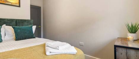 A neatly made bed with a yellow bedspread and white pillows, a small bedside table with a plant and candle, and a painting on the wall.
