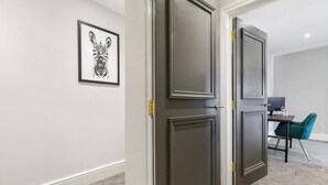 A hallway with two dark doors, a framed zebra artwork on the wall, and a desk with a computer and a teal chair. - WhiskAwayStays  Victoria House  Apartment 4 (Worcester)