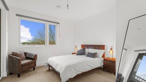 Ein Schlafzimmer mit einem großen Bett, einem Nachttisch, einem Sessel und einem Fenster mit Blick auf Gebäude und Bäume.