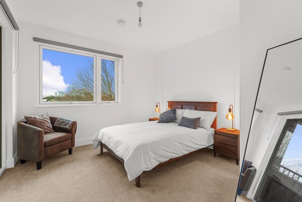 A bedroom with a large bed, a nightstand, a chair, and a window with a view of buildings and trees. - London - 1 Bed Apartment - Sleeps 2 - Long Stays (London)