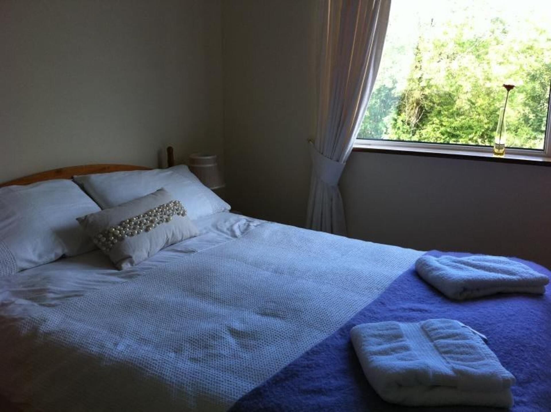 Een net opgemaakt bed met witte lakens en een blauwe deken, een raam met gordijnen, en uitzicht op groen.
