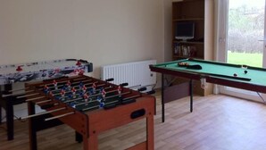 Una habitación con mesa de fútbol, mesa de billar, estantería y ventana con vistas a la vegetación.
