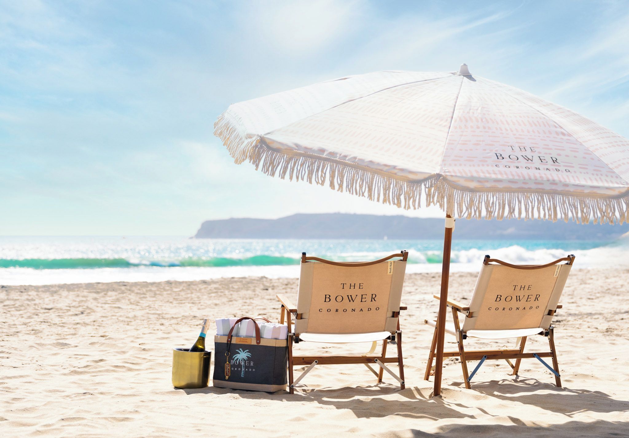 Twee strandstoelen met de branding 'The Bower Coronado', een strandparasol en een emmer met een fles champagne op een zandstrand met golven op de achtergrond.