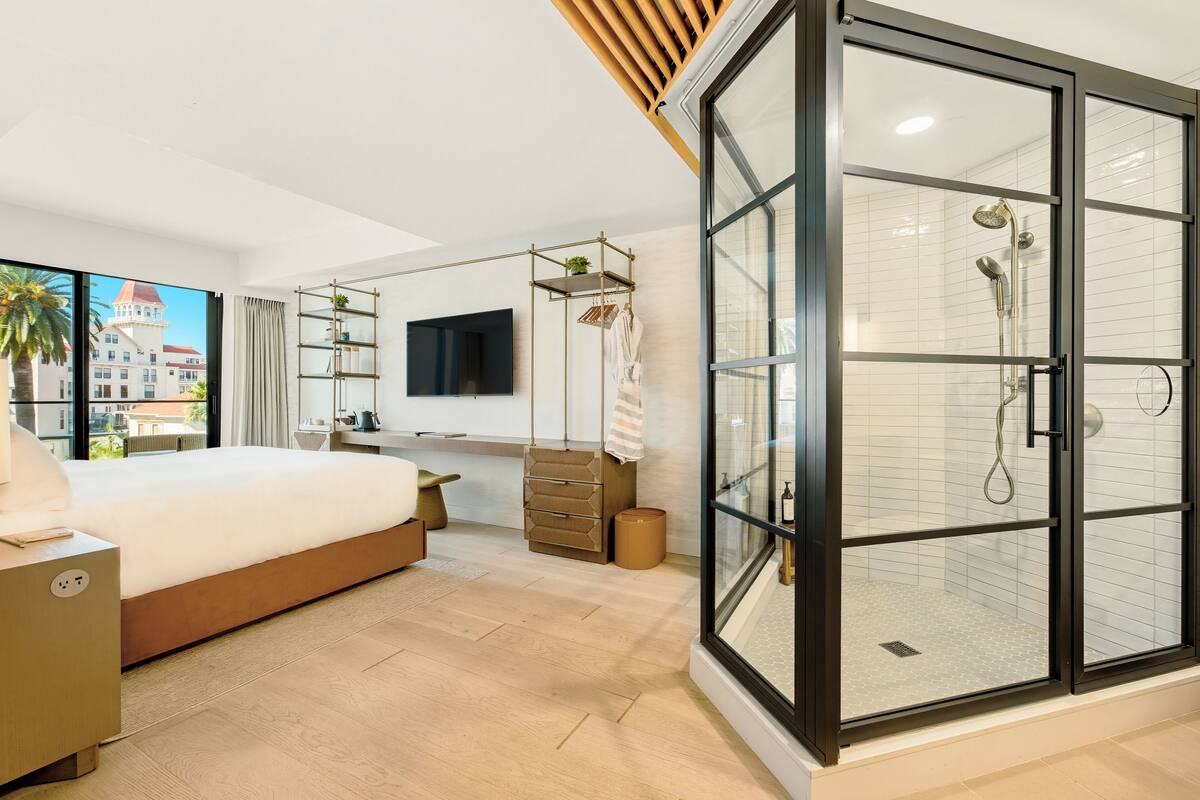 A modern bedroom with a large bed, a desk, a TV, a walk-in shower, and a city view.