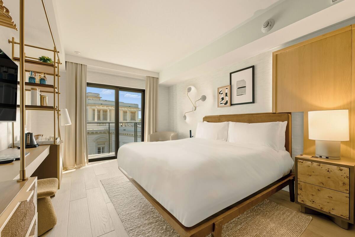 A hotel room with a large bed, a desk, a chair, a nightstand, and a view of the city.