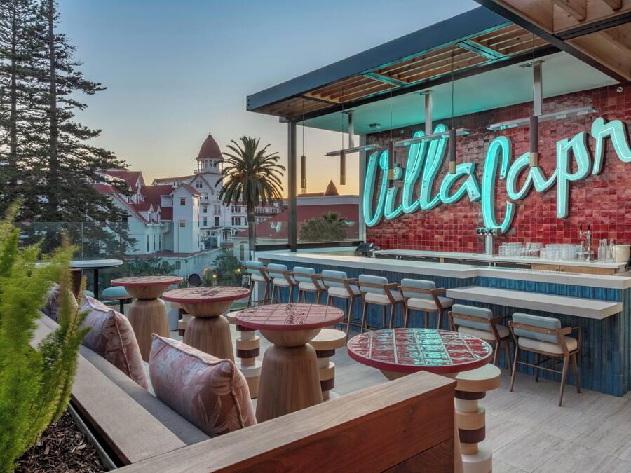 Un restaurant avec une enseigne au néon indiquant « Villa Capri », des tables et des chaises rondes disposées en terrasse, et une vue sur un bâtiment avec un toit rouge en arrière-plan.