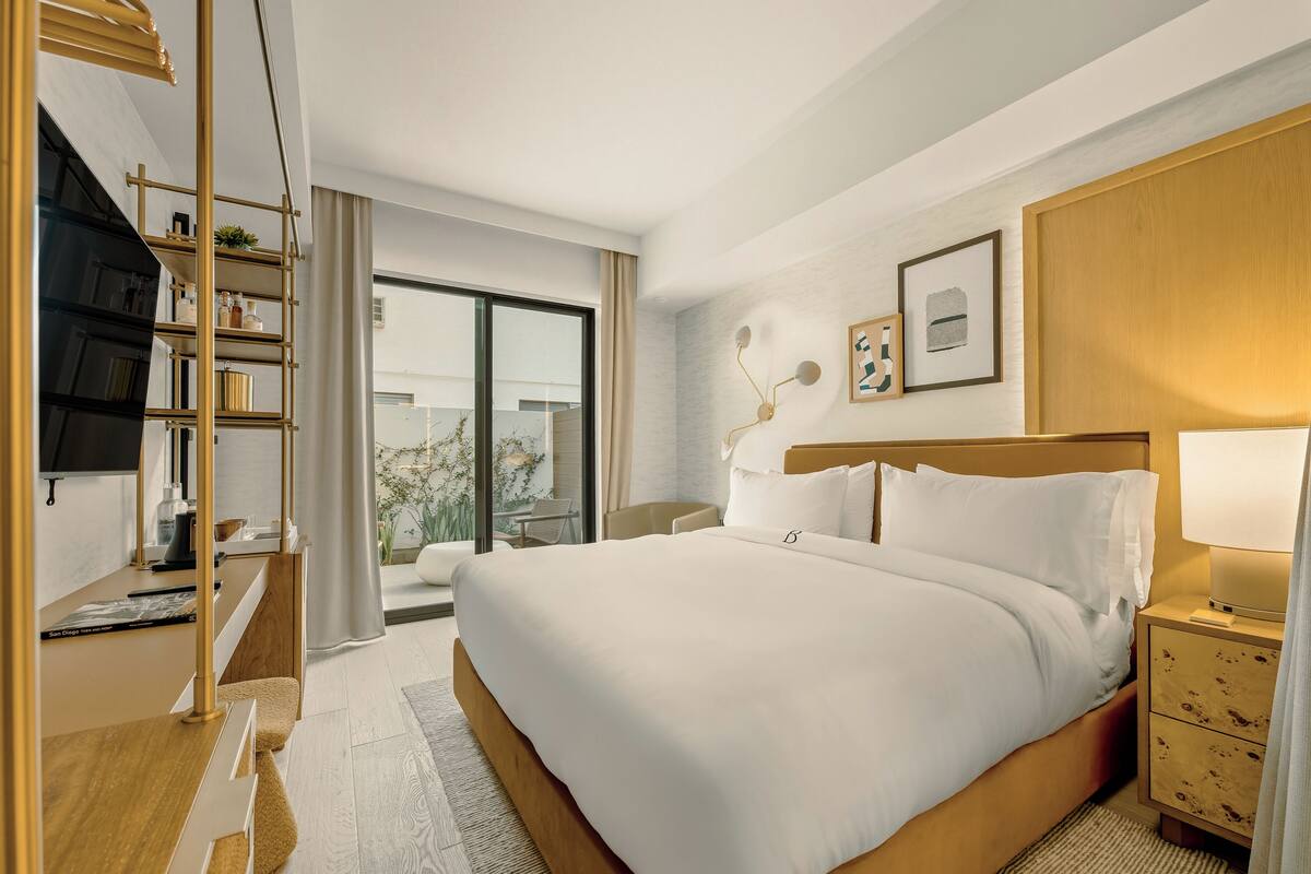 A hotel room with a large bed, a desk with a TV, a bookshelf, and a window with a view.