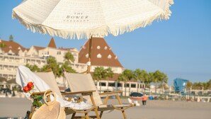 A beach chair under a beach umbrella with a rose and a bottle on the sand. - The Bower Coronado (Coronado)