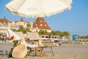 Une chaise de plage sous un parasol, avec une rose et une bouteille posées sur le sable.