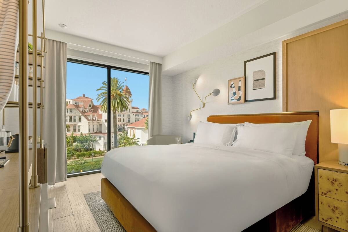 A hotel room with a large bed, a nightstand, a lamp, a window with a view of buildings and palm trees, and a door with curtains.